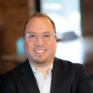 A picture of AJ DeLeón wearing glasses and a dark blazer smiling in a softly lit indoor setting.