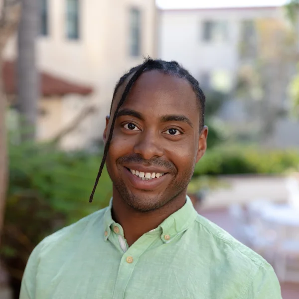 Smiling Aaron Fitzgerald in a light green button-up shirt standing outdoors with soft greenery and buildings in the background.