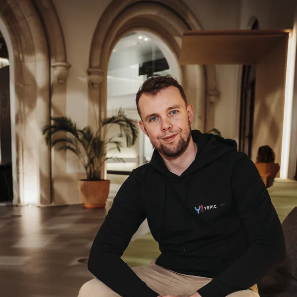 Smiling Aaron Jones in a black hoodie seated indoors, with arched doorways and potted plants in the background
