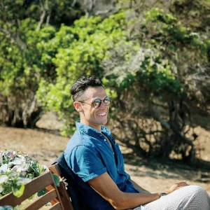 Adam Bent smiles while sitting on a bench in front of green trees.