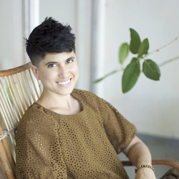 Alexandra Ballensweig smiling while seated in a wooden chair wearing a textured brown top indoors.
