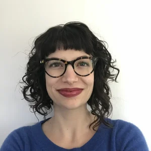 Alice Albrecht with short curly dark hair, glasses, and a blue sweater smiling in front of a white background.