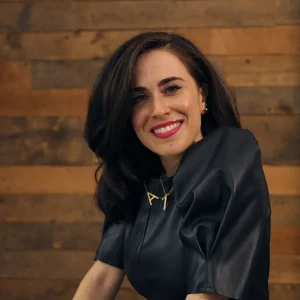 Smiling Alison Greenberg with dark hair wearing a black top and gold necklace, seated in front of a wooden wall