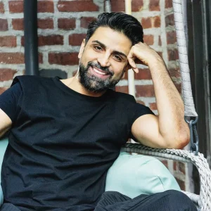 Amir Baradaran sitting and smiling on a cushioned chair against a brick wall backdrop.