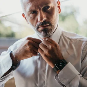 Armando Biondi tightening the collar of a white dress shirt, wearing a smartwatch, with a serious expression.