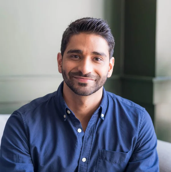 Asher Ismail smiling while seated indoors, wearing a blue button-down shirt.