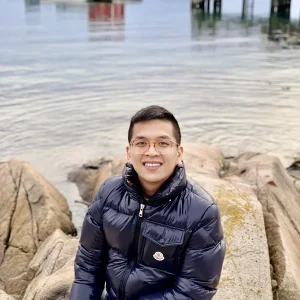Atom Nguyen in a dark puffer jacket smiling while sitting on rocks by the water.