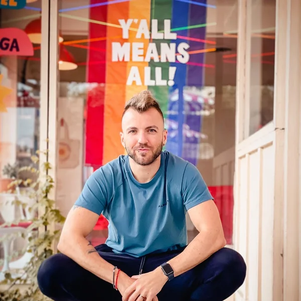 Aydian Dowling sitting outside a shop with a rainbow flag reading ‘Y’all Means All’ behind them.