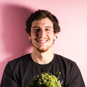 Bruno Sindicic smiling in front of a pink background while holding a large broccoli head.