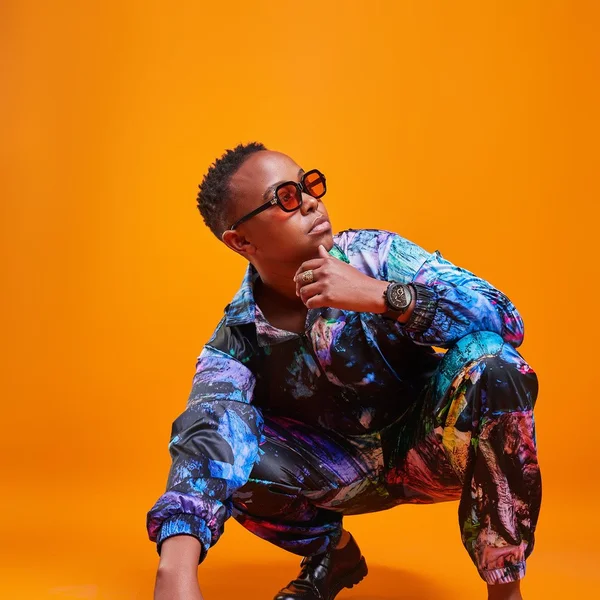 Chris Muriithi in colorful patterned clothing squatting confidently against a bright orange background.