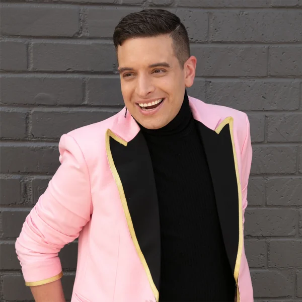 Damian Pelliccione smiling while wearing a pink blazer with gold trim against a gray brick wall.