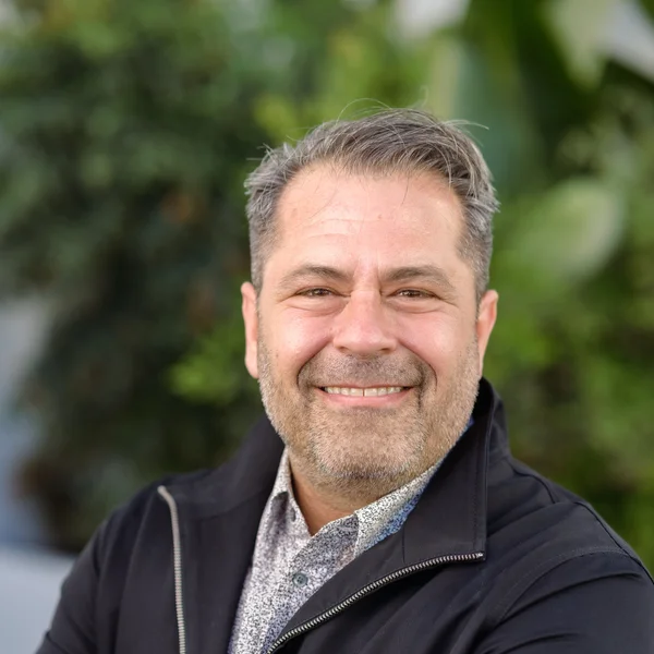 David Ehrich smiling warmly outdoors while wearing a dark jacket and patterned shirt.