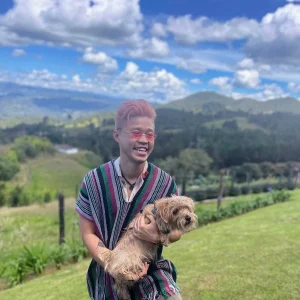 Edric Subur smiling outdoors while holding a small dog against a scenic mountain landscape.
