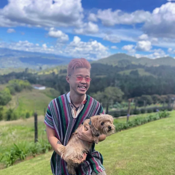 Edric Subur smiling outdoors while holding a small dog against a scenic mountain landscape.