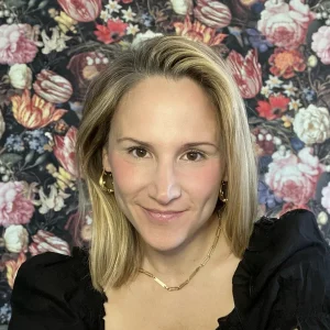 Emilie Poteat smiling in front of a dark floral patterned background while wearing gold jewelry.