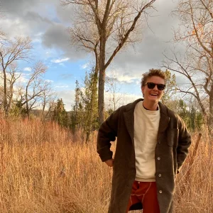 Smiling Finnegan Shepard in sunglasses standing in a field with tall grass and bare trees.