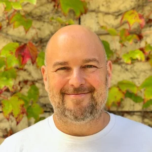 Smiling Gabe Zichermann standing outdoors in front of a wall covered with green and red leaves.