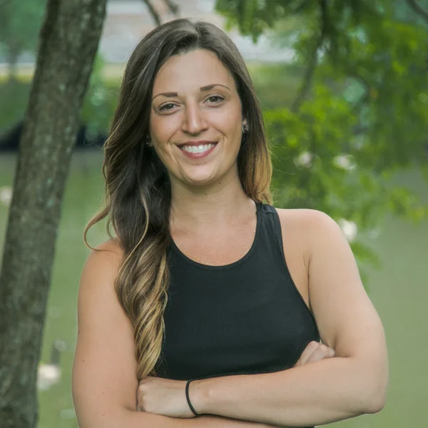 Gabriella Silva smiling outdoors with arms crossed in front of a tree and water.