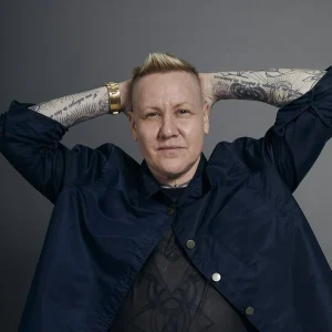 Grey Leifer with tattooed arms posed with hands behind head wearing a dark jacket.