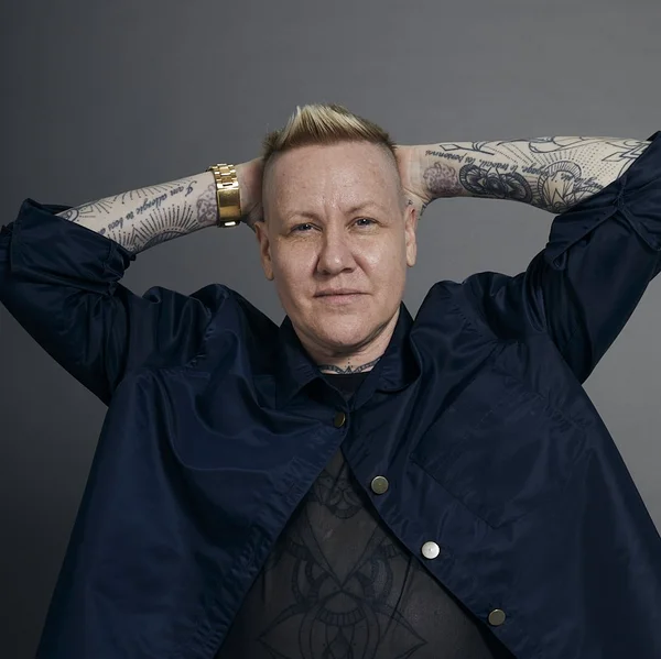 Grey Leifer with tattooed arms posed with hands behind head wearing a dark jacket.
