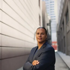 Heather Hiles in a dark suit standing in an alleyway with arms crossed, city buildings behind.