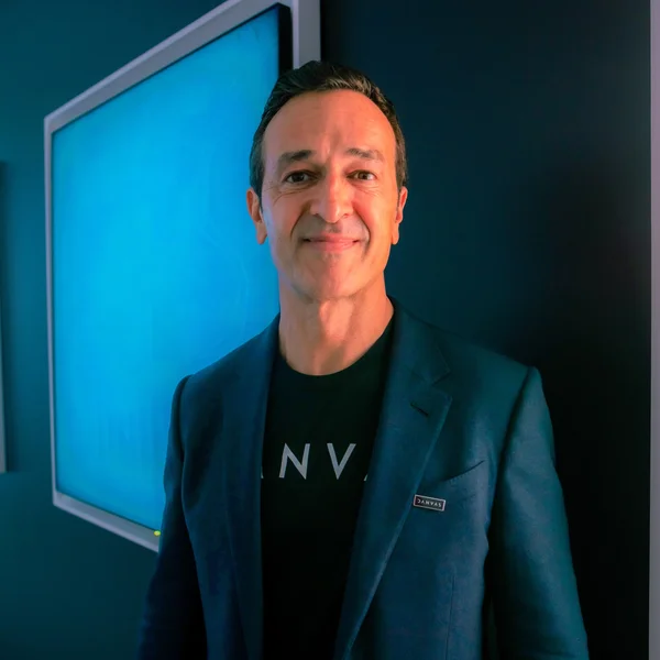Hernan Lopez in a dark blazer and T-shirt smiling while standing indoors beside a large blue-lit display screen.