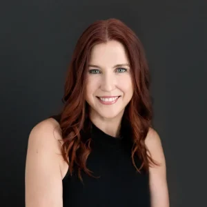 Irena Pereira with long wavy red hair smiling while posing against a dark neutral background.