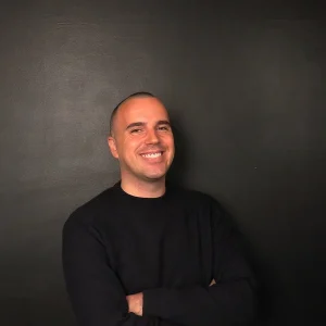 Ivan Kadic smiling with arms crossed while standing against a plain dark background.