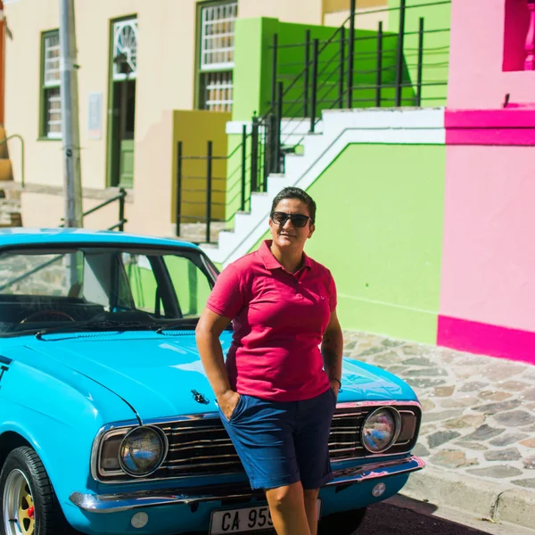 Janine Salame wearing sunglasses and a red polo shirt standing in front of a bright blue vintage car on a colorful street with pastel buildings
