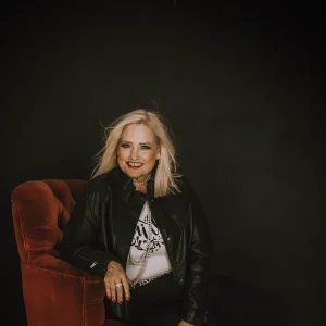 Jen Bolin with blonde hair smiling while seated in a red chair wearing a black jacket.