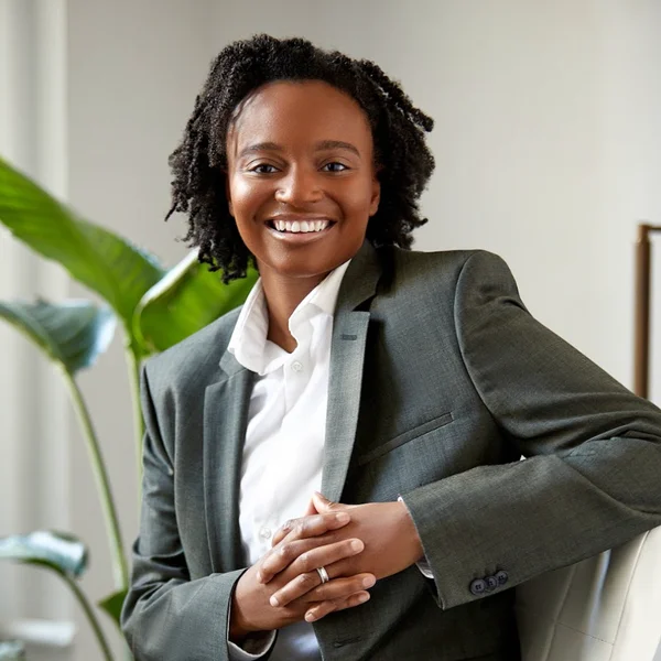 Smiling Jessica McGlory in a gray suit jacket leaning on a chair with plants behind.
