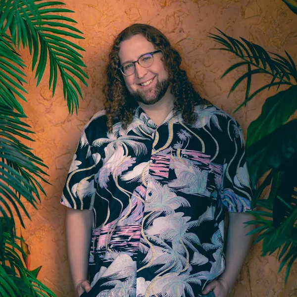 Jo Graylock with long curly hair and glasses wearing a patterned shirt, standing between tropical plants against an orange wall.