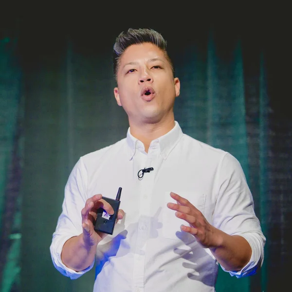 Jonathan TranPham speaking onstage in a white shirt while holding a small wireless microphone device.