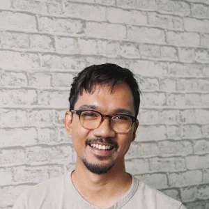 Joseph Undaloc smiling in glasses while standing against a light gray brick-patterned wall.