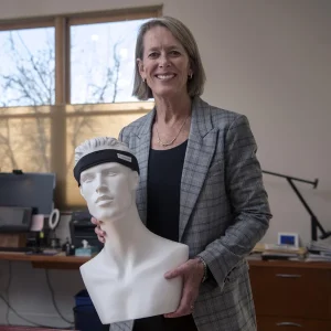 Smiling Karen Crow in blazer holding a white mannequin head wearing a black headband device.
