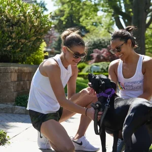 Kathryn Ruhno-McCallum sitting outdoors on a sunny day, smiling and petting a black dog wearing a purple collar.