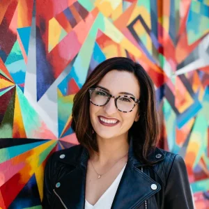 Kayla Glanville smiling in glasses and a leather jacket standing before a colorful mural.