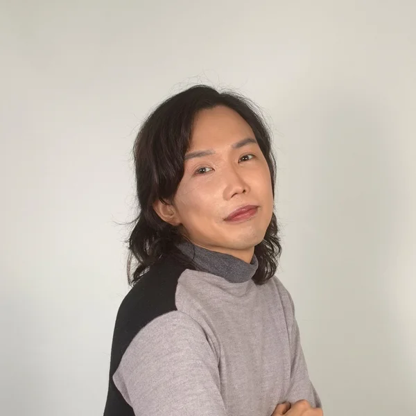 Kenji-Travis Yamazaki posing with folded arms in a gray sweater against a plain light background.
