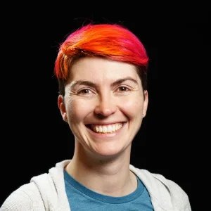 Smiling Leigh Honeywell with short bright orange-and-pink hair against a black background.