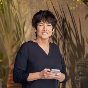 Lil Roberts holding a smartphone and smiling while standing in front of leafy shadows.