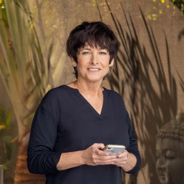 Lil Roberts holding a smartphone and smiling while standing in front of leafy shadows.