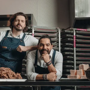 Luis Gramajo and Hans Schrei in aprons posing in a kitchen beside large trays and blocks of dough.
