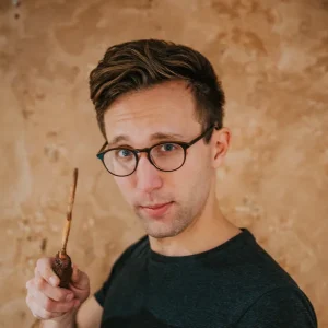 Matthew Cortland holding a small wooden object and looking toward the camera against a tan wall.