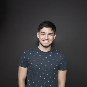 Smiling Michael Sayman standing against a dark background wearing a patterned T-shirt.