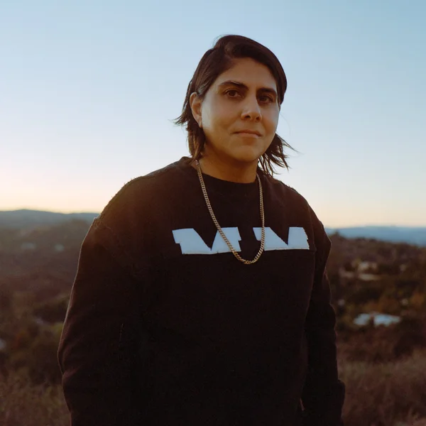 Moj Mahdara standing outdoors at sunset, wearing a black sweatshirt and gold chain, with hills in the background.