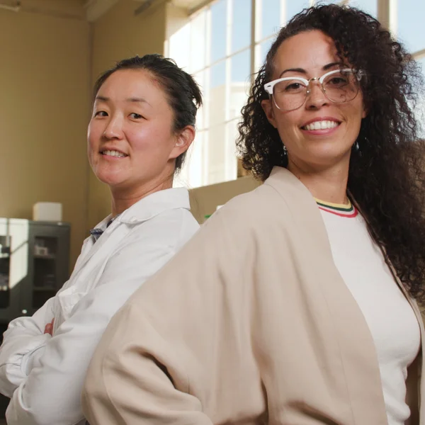 Nieves Martínez-Marshall and Michelle Lu stand back to back smiling in a bright workspace.