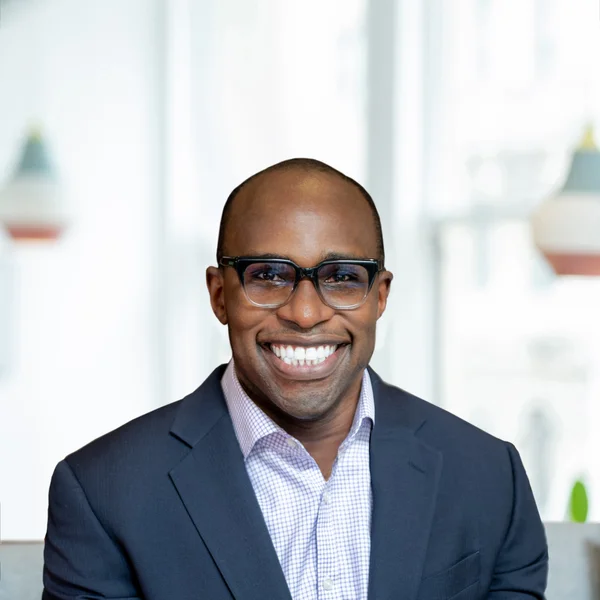 Smiling Perrin Quarshie in glasses wearing a suit jacket and patterned shirt indoors.