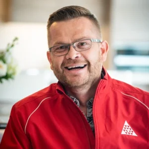 Raymond Watt wearing glasses and a red jacket smiling in a bright indoor setting.