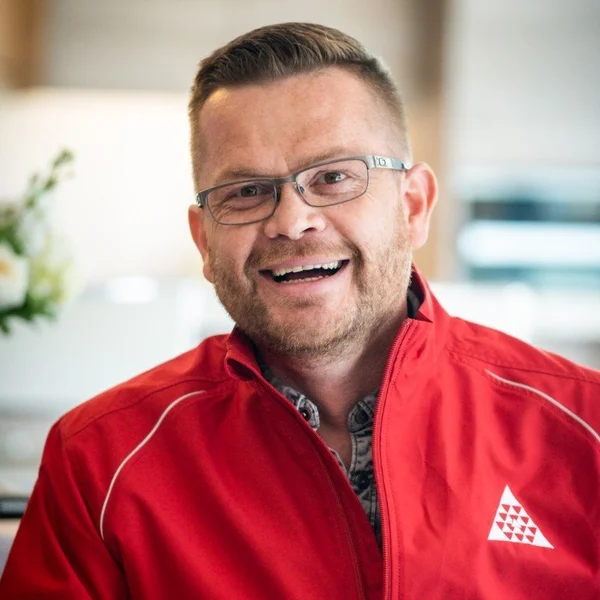 Raymond Watt wearing glasses and a red jacket smiling in a bright indoor setting.