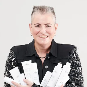 Smiling Rochelle Weitzner in a black jacket holding multiple white skincare product bottles.
